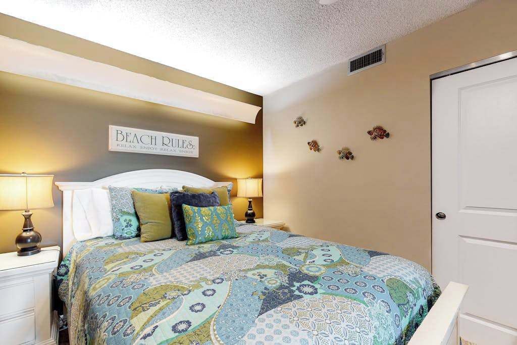 Une chambre à coucher comprenant un lit, deux tables de chevet, une commode, un panneau fixé au mur et une porte.