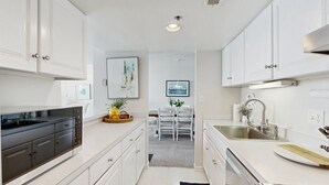 A modern kitchen with white cabinetry, a dining area with a table and chairs, and a view of the cityscape through the window. - Sea Colony Ocean 7th floor condo w/ basketball court, pool, and elevator (Bethany Beach)