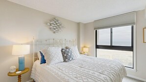 A bedroom with a bed, two bedside lamps, a decorative wall piece, and a large window. - Sea Colony Ocean 7th floor condo w/ basketball court, pool, and elevator (Bethany Beach)