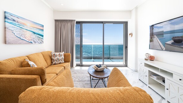 A modern living room with a sofa, a coffee table, and a TV. There is a large window with a view of the sea. - Beachfront 2BR Condo Pool Ocean Views Resort (Puerto Peñasco)