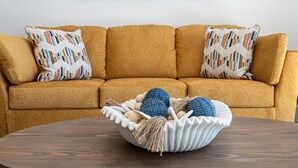 A mustard-colored sofa with patterned pillows, a woven basket on a wooden table, and a beach-themed painting on the wall. - Beachfront 2BR Condo Pool Ocean Views Resort (Puerto Peñasco)