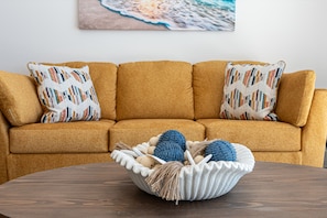 A mustard-colored sofa with patterned pillows, a woven basket on a wooden table, and a beach-themed painting on the wall. - Beachfront 2BR Condo Pool Ocean Views Resort (Puerto Peñasco)