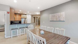A dining area with a wooden table and white chairs, a kitchen with wooden cabinets, and a gray wall with a painting. - Bayside Resort 4th floor condo w/ gym, tennis court, and free WiFi (Selbyville)