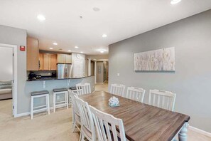A dining area with a wooden table and white chairs, a kitchen with wooden cabinets, and a gray wall with a painting. - Bayside Resort 4th floor condo w/ gym, tennis court, and free WiFi (Selbyville)