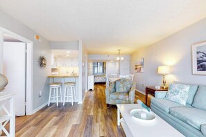 A modern living room with a sofa, a dining table, and a kitchen area in the background. - Sea Colony Ocean 6th floor condo w/ tennis court, balcony, and shared sauna (Bethany Beach)
