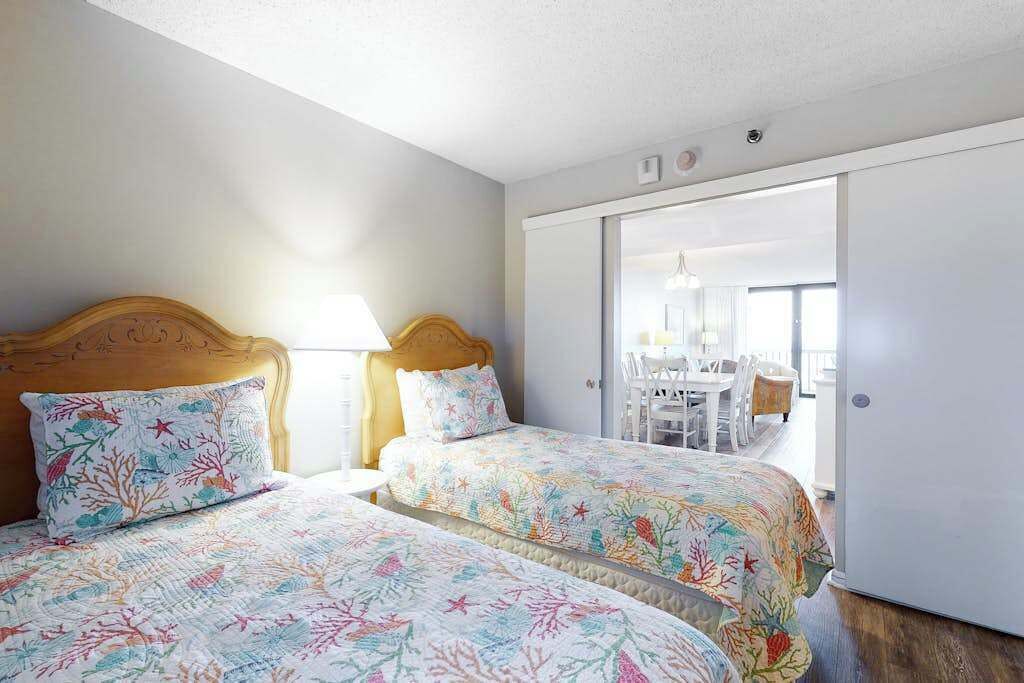 Une chambre avec deux lits, une tête de lit en bois, une table de chevet et une porte coulissante donnant sur un coin repas.