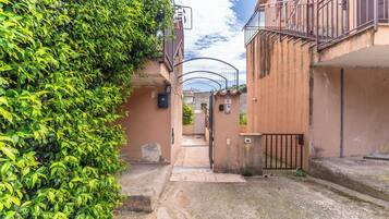 A narrow alleyway with a gate, a building with a balcony, and a hedge on the left.