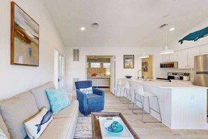 Una sala moderna con un sofá, un sillón azul, una mesa de centro y una zona de cocina con barra y armarios blancos.