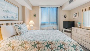 Une chambre d’hôtel avec un lit, une télévision, une commode, une fenêtre donnant sur la plage et un tableau au mur.