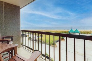 Ein Balkon mit Blick auf den Strand, ein Holztisch und ein gebäudeähnliche Struktur mit einem grünen Dach im Hintergrund.