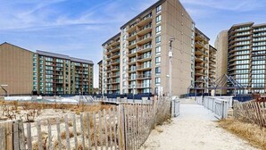 Ein modernes Mehrfamilienhaus mit mehreren Etagen und Balkonen, ein sandiger Weg führt zum Eingang.