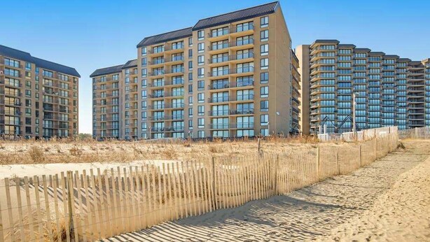 Una fila di edifici residenziali a più piani con numerosi balconi, una spiaggia sabbiosa in primo piano e un cielo azzurro limpido.