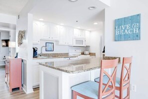 A modern kitchen with white cabinets, a granite countertop, and a central island with bar stools. - Sea Colony Ocean 5th floor condo w/ shared sauna, elevator, and pool (Bethany Beach)