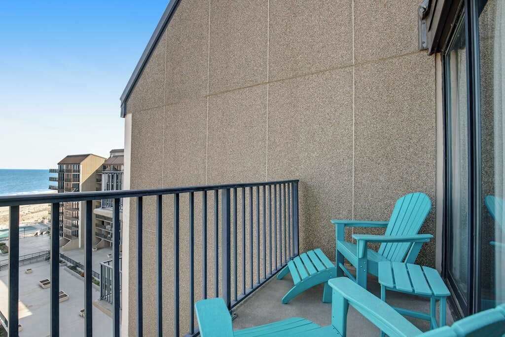 Een balkon met turquoise Adirondack stoelen, uitzicht op het strand en een gebouw met een structurele gevel.