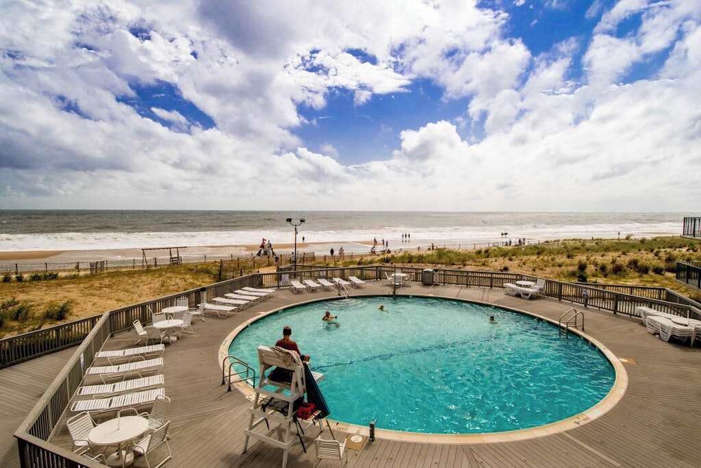 A poolside area with a view of the beach, lounge chairs, and a wooden deck.