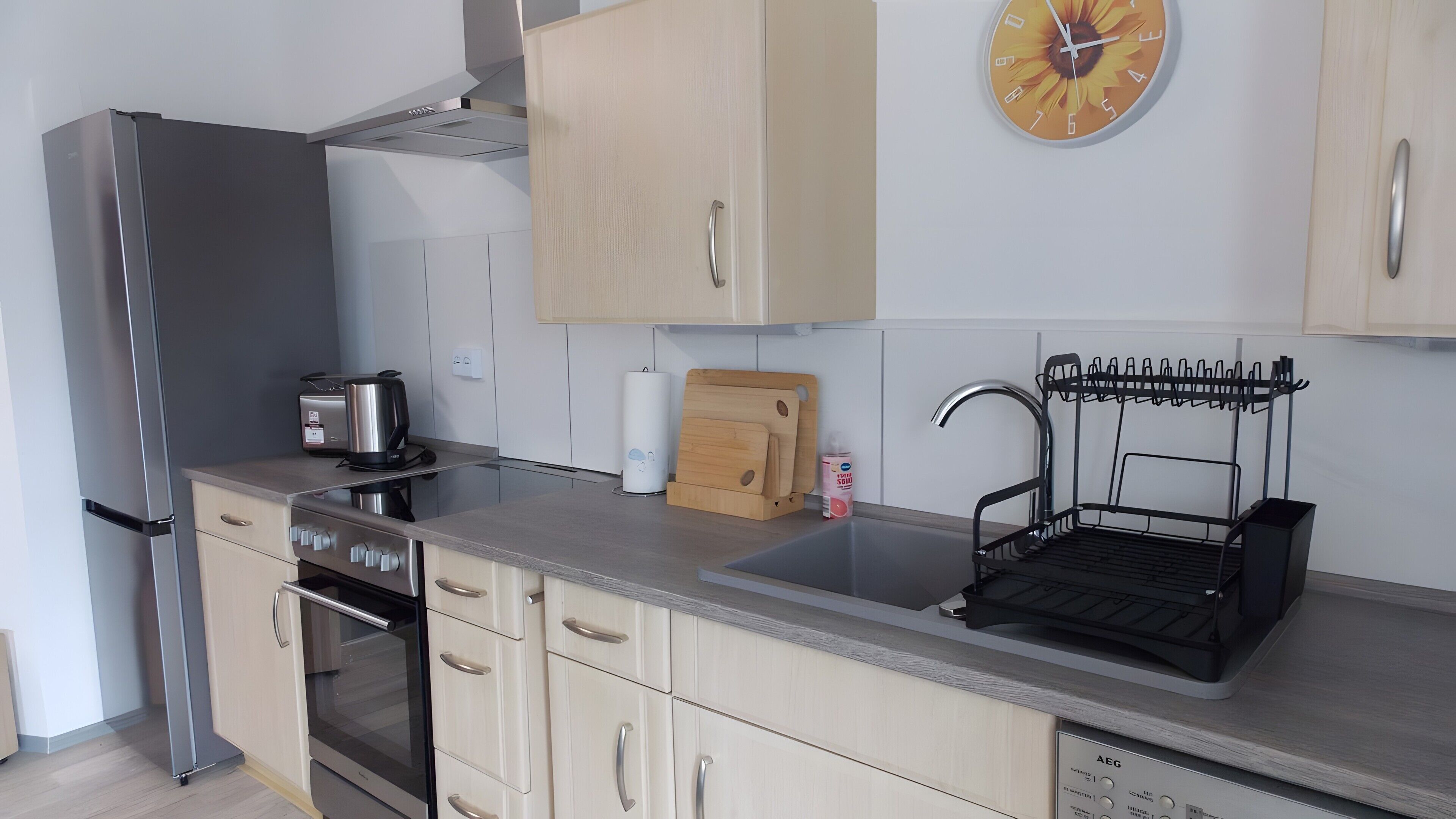 Eine moderne Küche mit einem Edelstahl-Kühlschrank, einem eingebauten Backofen, einer Spüle mit Abtropfgestell und einer an der Wand montierten Uhr.