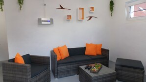 A modern living room with wicker furniture, a central sofa with orange cushions, a coffee table with decorative items, and wall-mounted decor including a thermostat and bird sculptures.