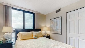 Habitación de hotel con una cama grande, dos mesitas de noche, una ventana con cortinas, una puerta y un cuadro enmarcado en la pared.
