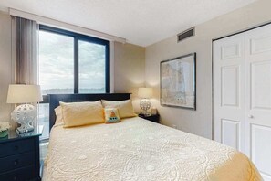 Een hotelkamer met een groot bed, twee nachtkastjes, een raam met gordijnen, een deur en een lijst met een afbeelding aan de muur.