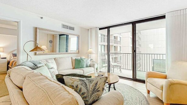 Un salón moderno con sofá, sillón y una mesa de centro redonda. Hay un espejo grande en la pared, una ventana con vistas al exterior y un balcón con barandilla.