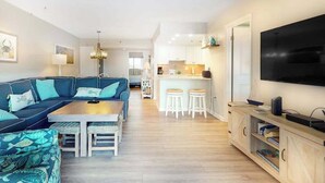 A modern living room with a sofa, coffee table, and bar stools. There is a kitchen in the background with white cabinets and a flat-screen TV mounted on the wall. - Sea Colony Ocean 5th floor condo w/ tennis court, pool, and elevator (Bethany Beach)