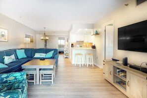 Ein modernes Wohnzimmer mit einer Couch, einem Couchtisch und Barhocker. Im Hintergrund ist eine Küche mit weißen Schränken und einem an der Wand befestigten Flachbildfernseher zu sehen.