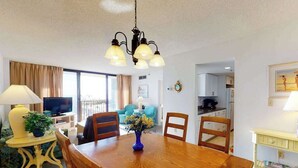 A dining area with a wooden table, chairs, a vase with flowers, a television, and a kitchen in the background. - Sea Colony Ocean 2nd floor condo with sauna (Bethany Beach)