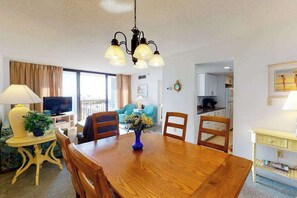 Ein Essbereich mit Holztisch, Stühlen, einer Vase mit Blumen, einem Fernseher und einer Küche im Hintergrund.