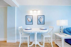 Un comedor con una mesa y sillas blancas, dos cuadros en la pared y una lámpara sobre una mesita auxiliar.