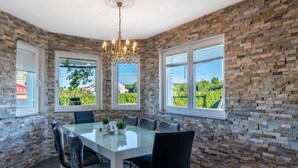 Un espace repas avec une table en verre, des chaises noires, un lustre, des murs en pierre et de grandes fenêtres donnant sur un paysage verdoyant.