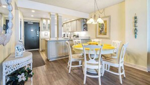 A modern kitchen with white cabinets, a central island, and a dining area with white chairs and a yellow tablecloth. - Sea Colony Ocean 6th floor condo w/ pool, gym, and free WiFi (Bethany Beach)