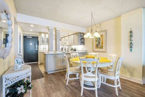 A modern kitchen with white cabinets, a central island, and a dining area with white chairs and a yellow tablecloth. - Sea Colony Ocean 6th floor condo w/ pool, gym, and free WiFi (Bethany Beach)