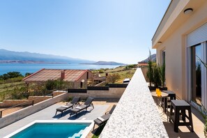 Uma área à beira da piscina com espreguiçadeiras, uma mesa e vista para o mar e as montanhas.