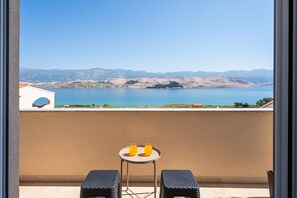 A balcony with a view of a lake and mountains, two chairs, and a table with two glasses of orange juice.