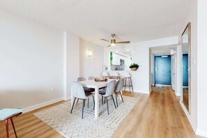 Un espace repas moderne avec une table blanche et des chaises grises, un ventilateur de plafond et une cuisine en arrière-plan.