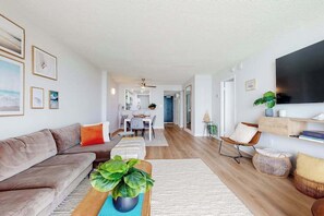 A modern living room with a sofa, coffee table, and a TV. There is a dining area in the background. - Sea Colony Ocean 7th floor condo with indoor/outdoor pools, tennis, & gym access (Bethany Beach)