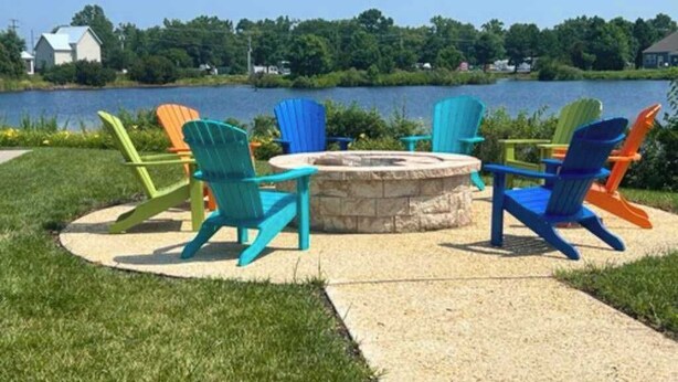 Een rond zitgedeelte met Adirondack-stoelen rond een vuurkorf van steen, gelegen op een betonnen terras met uitzicht op een meer.