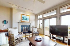 Una sala de estar con muebles de mimbre, chimenea, televisor y ventanas amplias.
