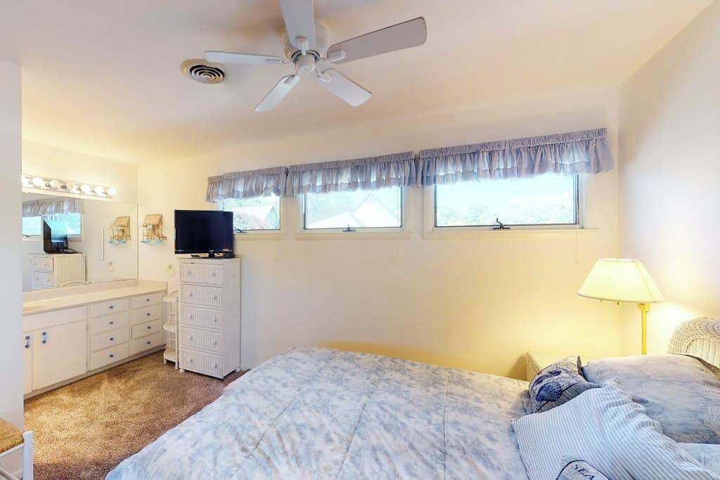 Ein Schlafzimmer mit Bett, Kommode, Fernseher, Deckenventilator und einem Fenster mit Vorhängen.