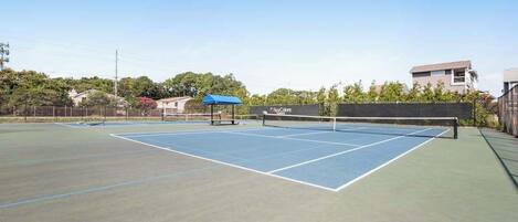 An outdoor tennis court with a net, surrounded by a low fence, and buildings in the background.
