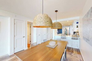 Ein moderner Essbereich mit einem Holztisch, zwei gewebten Pendelleuchten, weißen Stühlen und einer Küche im Hintergrund.