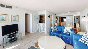 A living room with a TV, a blue sofa, a round coffee table, and a kitchen in the background. - Sea Colony Ocean 7th floor condo w/ gym, balcony, and tennis court (Bethany Beach)
