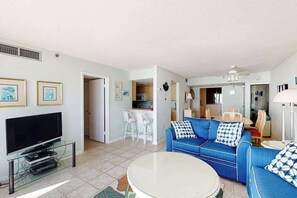 Ein Wohnzimmer mit Fernseher, einem blauen Sofa, einem runden Couchtisch und einer Küche im Hintergrund.