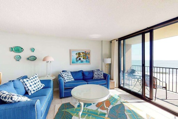 Una habitación con un sofá azul, una mesa redonda, un cuadro y vistas al mar a través de amplios ventanales.