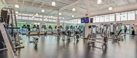 Un gimnasio amplio con diversas máquinas de ejercicio, ventanas grandes y un techo alto.