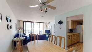A dining area with a wooden table and chairs, a ceiling fan, and a kitchen visible through an open doorway. - Sea Colony Ocean 7th floor condo w/ gym, balcony, and tennis court (Bethany Beach)