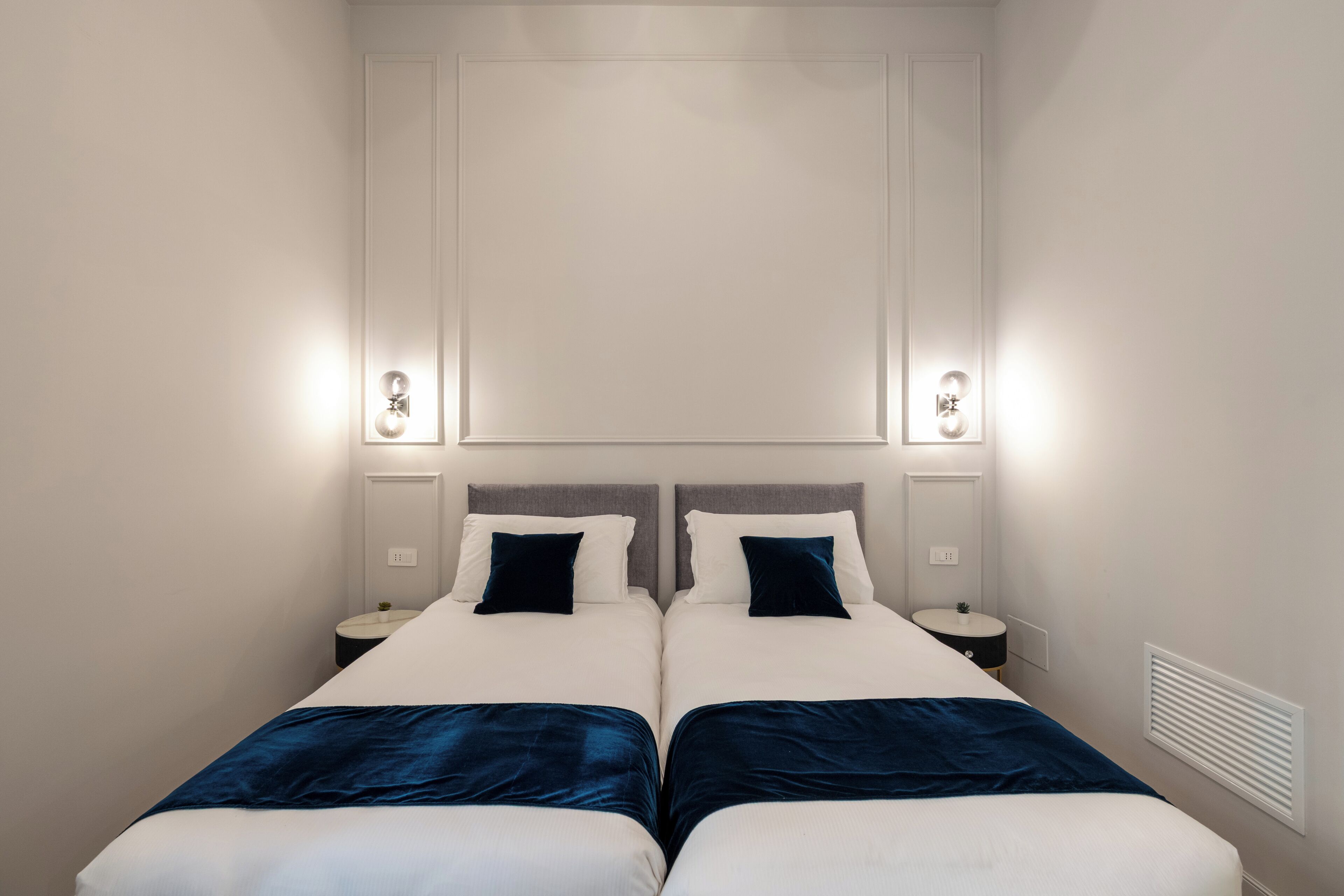 A double bed with two nightstands, each with a lamp, in a room with a plain wall and a vent.