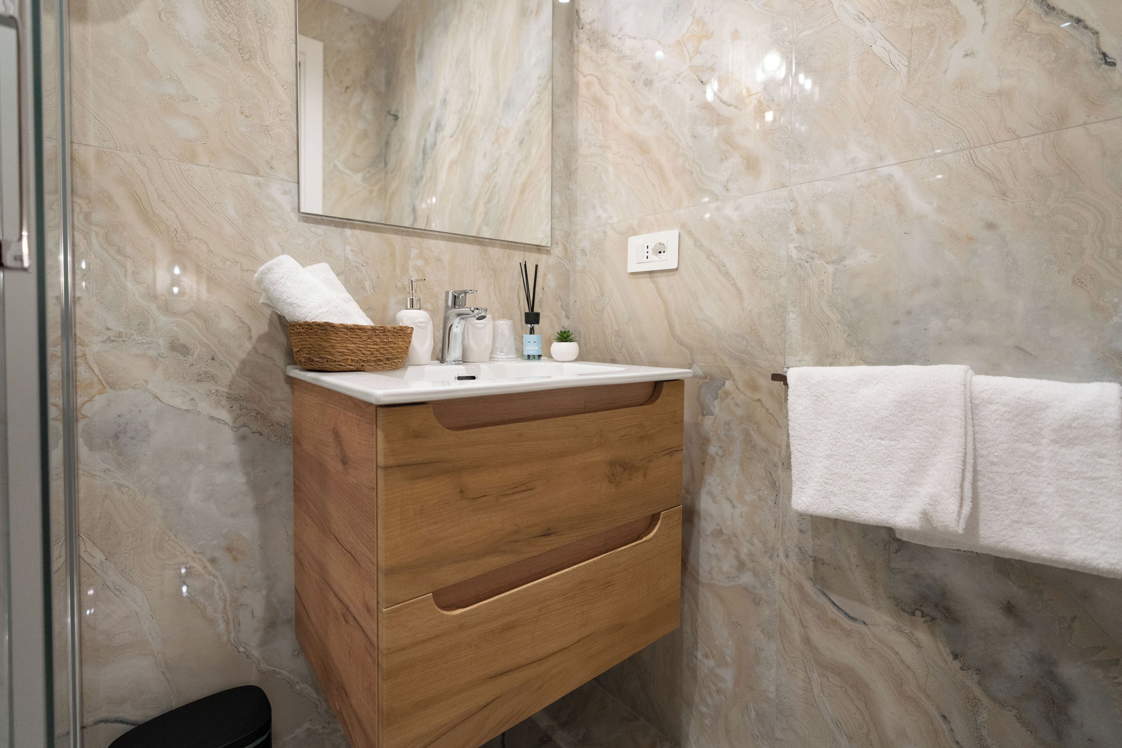Un baño con pared de mármol, tocador de madera, espejo, toallero y cesta con toallas.