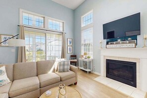 A living room with a sofa, fireplace, TV, and large windows. - Bayside Resort dog-friendly townhouse w/ tennis court access, WiFi, and balcony (Selbyville)