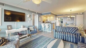 Un salon comprenant un canapé, des fauteuils, un téléviseur, un ventilateur de plafond et un coin cuisine.